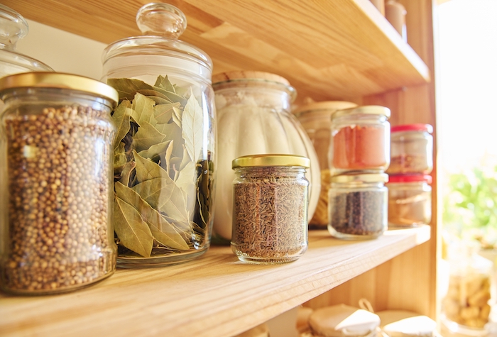 Various glass jars filled with spices on a wood shelf in a warmly lit pantry.