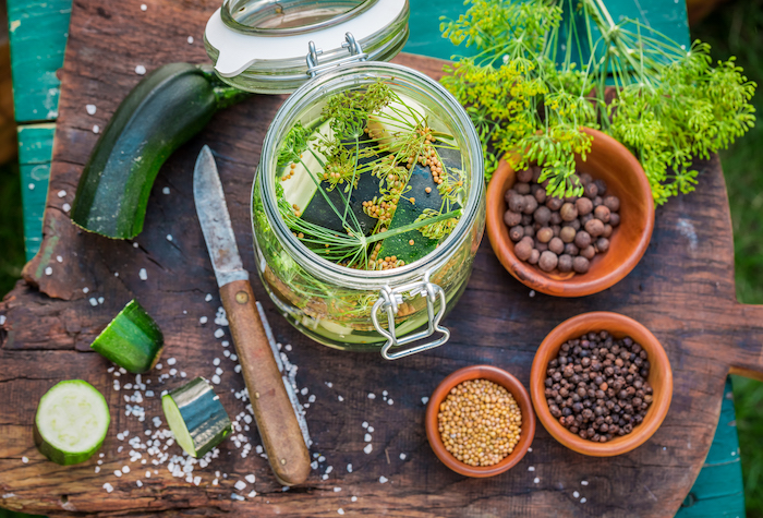 A dark wood cutting board on a green wood table outside. On the cutting board is a jar filled with spices and vegetables with a knife.