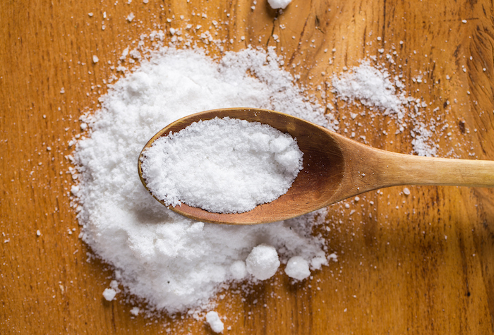 A wooden spoon full of table salt hovering above a brown kitchen table with a pile of table salt in the middle.