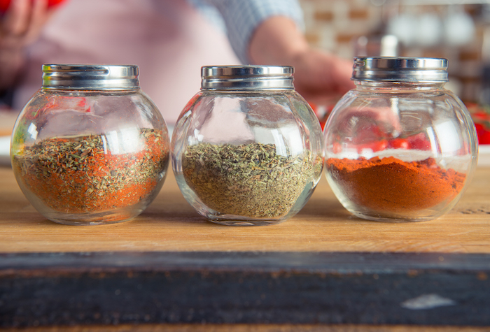 Three round glass jars filled with spices on a wood countertop and a baking woman in the background.