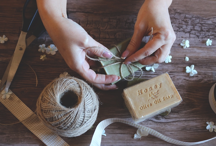 Person tying a bow with twine around a green, homemade bar of soap with scissors, ribbon, a ball of twine, and another bar of soap on the table.