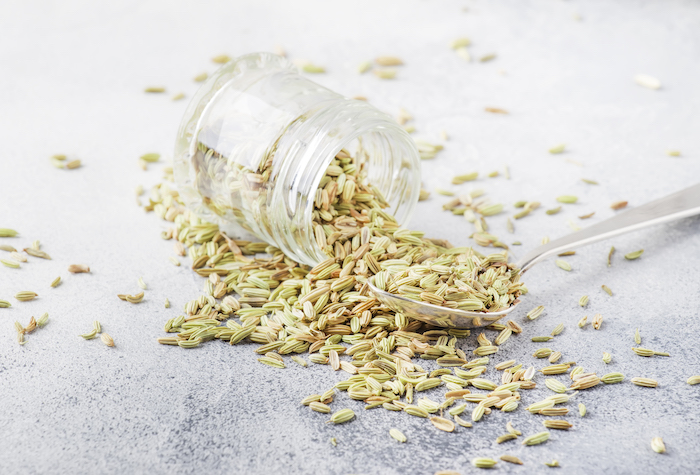 Fennel seeds in a glass jar on its side poured out onto a metal spoon and a table.